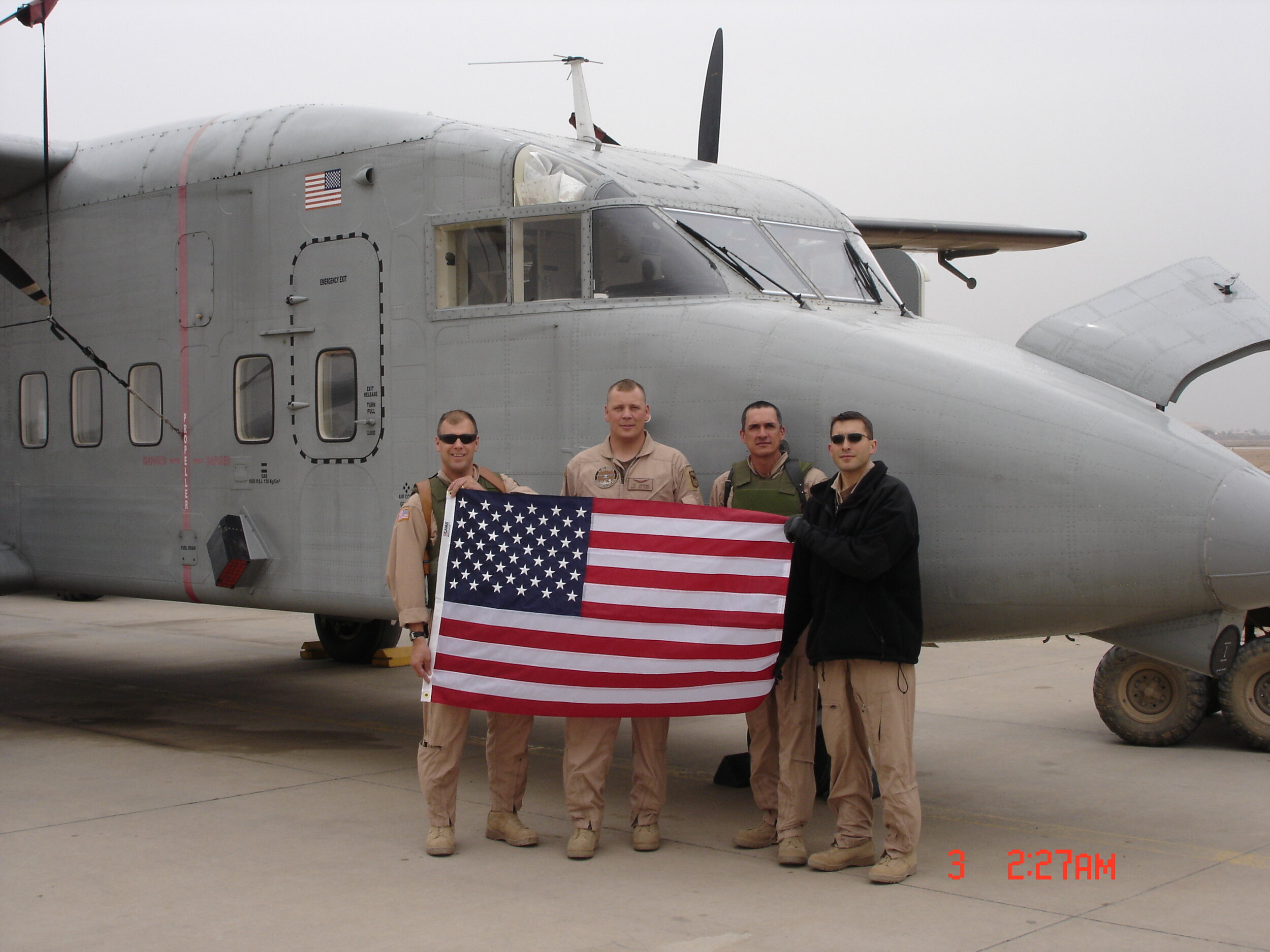 Southern+Sunny+Acres,+Texas+-+613E06CD-CF2B-410C-B8CC-50D80D9D3261 Four military personnel stand in front of a gray aircraft, holding an American flag. The image showcases camaraderie and patriotism, with the aircraft featuring military insignia and an American flag on its tail. The setting appears to be an airbase, emphasizing a military operation.